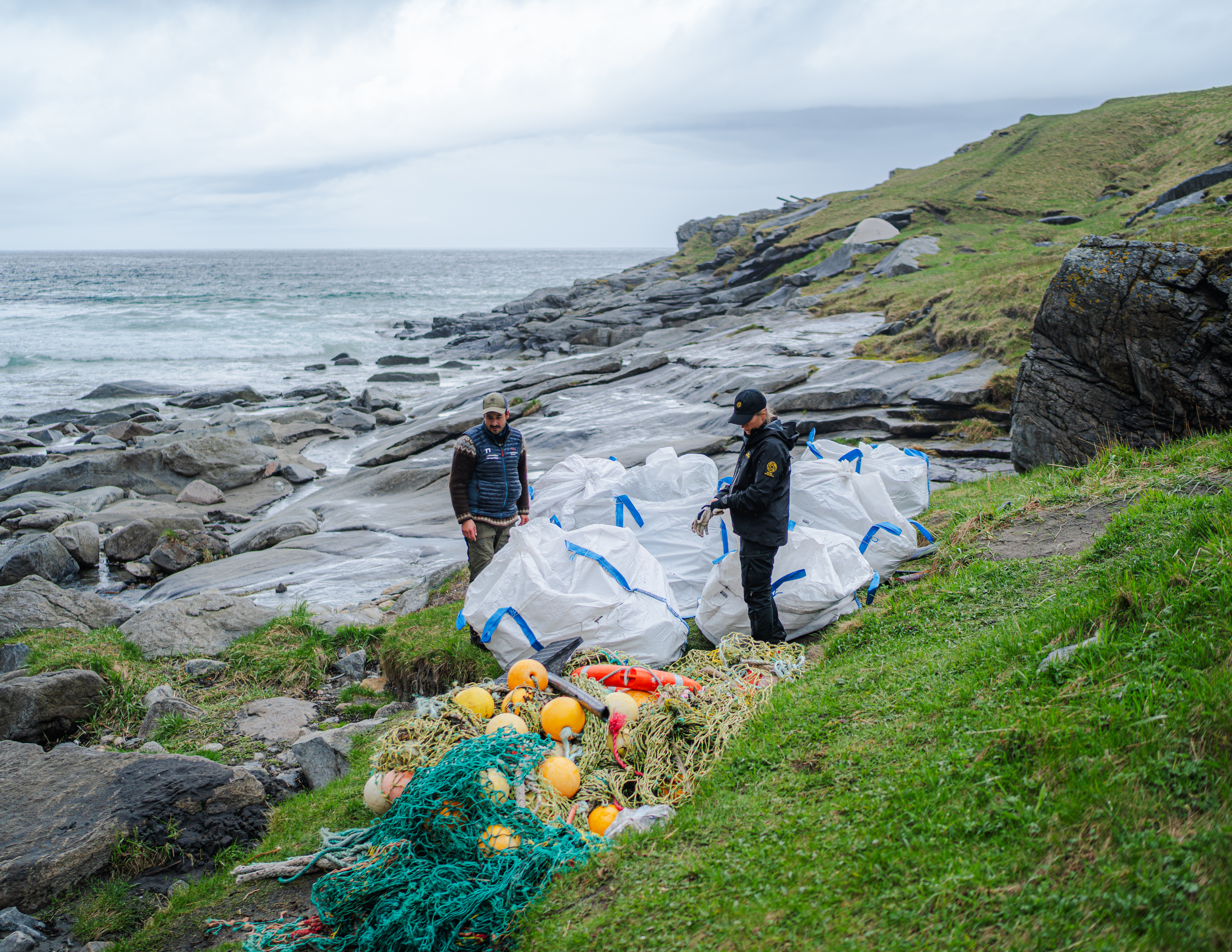 To personer pakker marint avfall ned i store kubiksekker. I bakgrunnen ses sandstranden Kvalvika og spisse fjell.